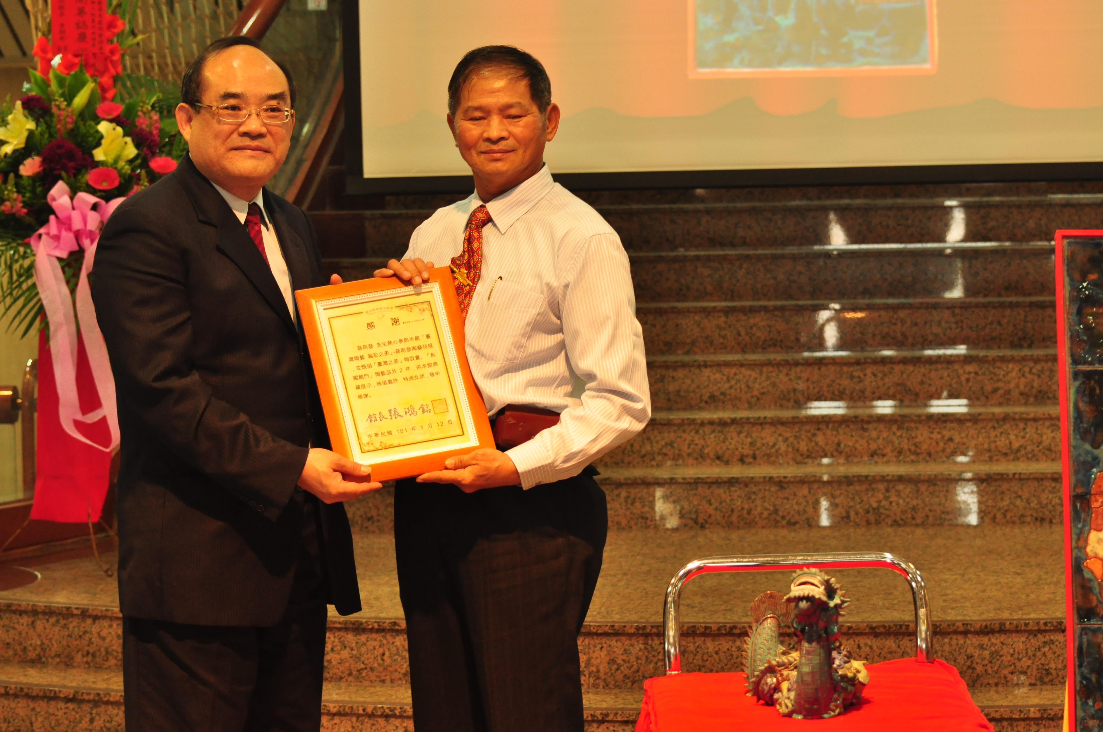 本館張館長鴻銘(左)致贈蔣再發先生(右)感謝狀，前為蔣先生捐贈之「魚躍龍門」陶藝品